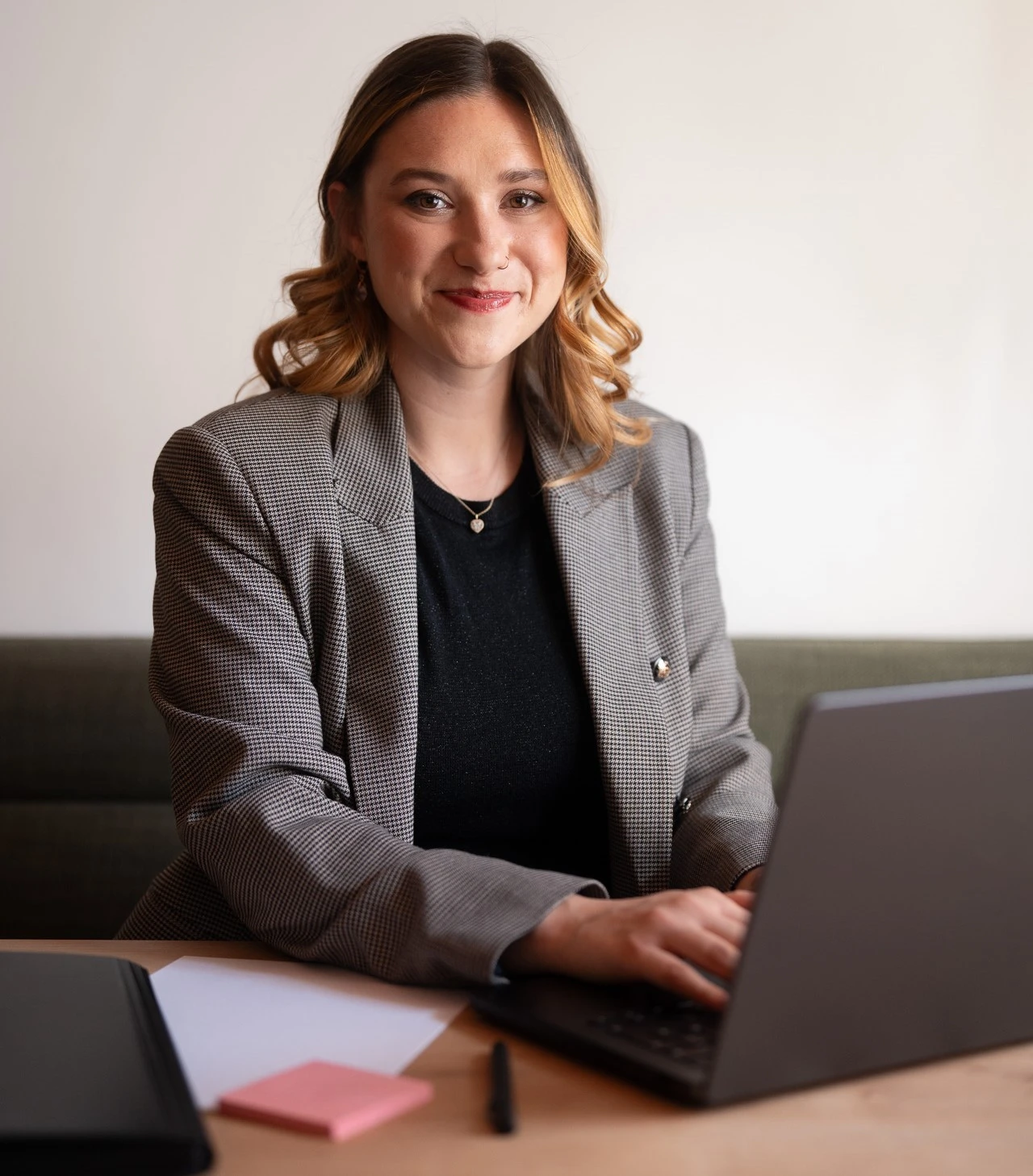 Lucia berät KMU, Selbstständige und Gründerinnen bei der Erstellung und Optimierung ihrer Homepage. Sie sitzt vor einem Schreibtisch vor ihr ein geöffneter Laptop und ein Notizblock. Sie tippt auf dem Laptop sieht aber in die Kamera. Ihre haare sind offen und sie trägt ein grau-karierte Sakko. Sie hat ein herzförmiges bis ovales Gesicht und braune lockige Haare.