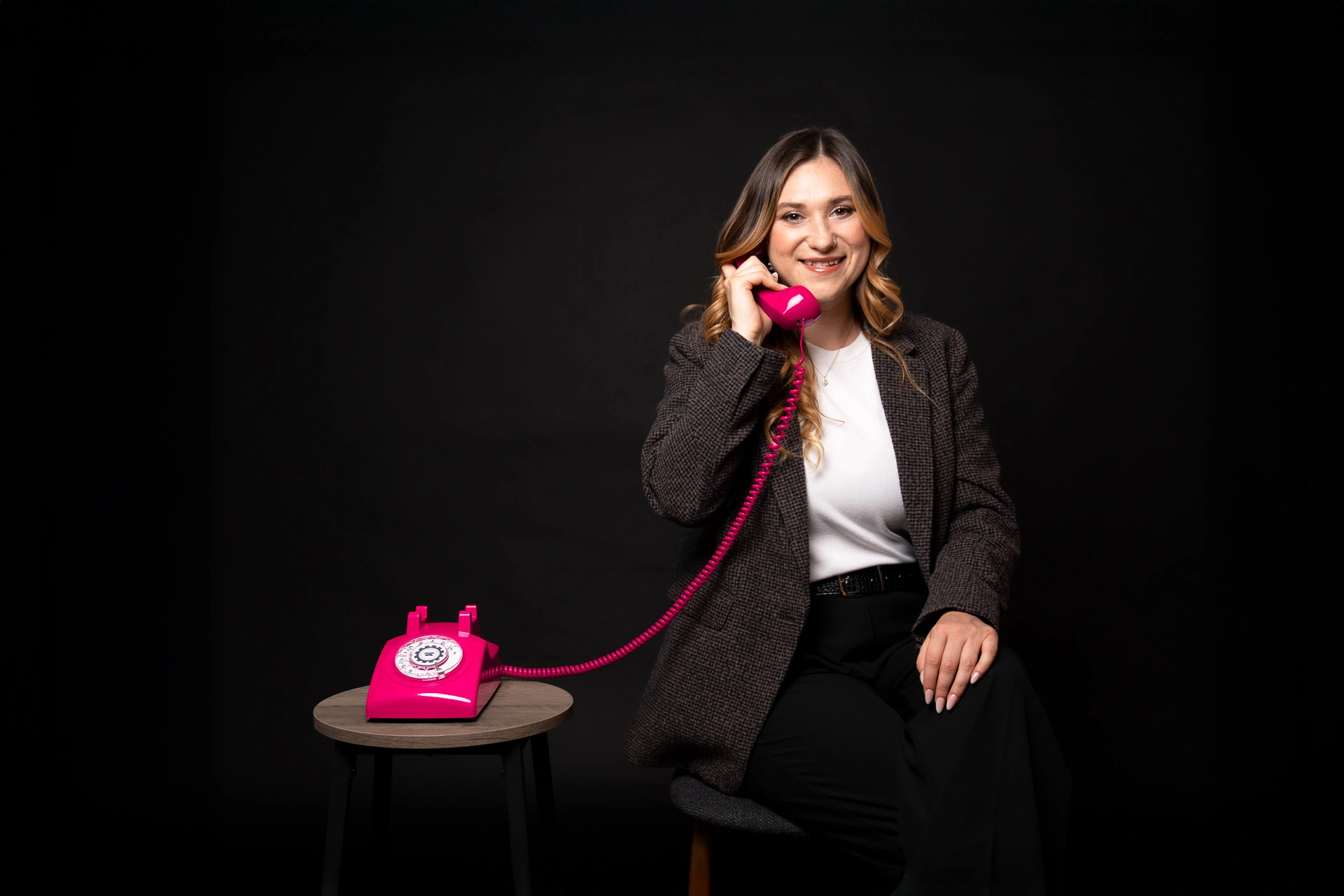 Lucia, PR-Beraterin bei Better Call Lucy, unterstützt Unternehmen bei Pressearbeit und Werbeschaltungen. Sie sitzt auf einem Stuhl neben einem kleinen Tisch auf dem ein pinkes Retro-Telefon mit Wahlscheibe liegt. Eine lange Telefonschnur führt den Hörer zu ihrem Ohr. Sie trägt ein legeres Business-Outfit vor schwarzem Hintergrund.