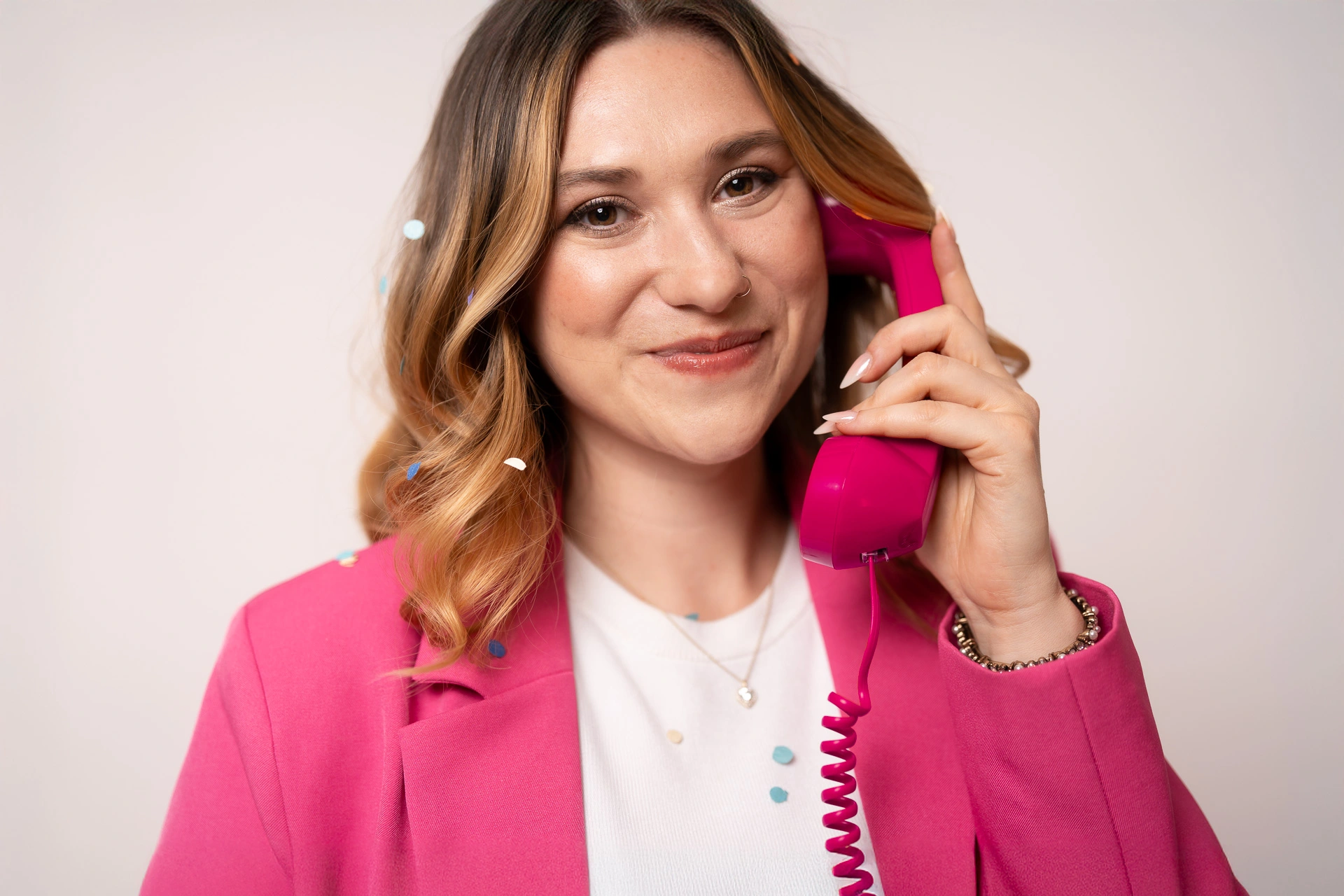 Lucia berät Unternehmen dabei einem erfolgreiche Social-Media-Strategie aufzubauen. Sie hält einen Hörer eines pinken Schnur-Telefons an ihr Ohr. Sie trägt ein pinkes Sakko und ein weißes T-Shirt sie lächelt in die Kamera. Sie trägt ihre braun-blonden Haare offen und in Locken.